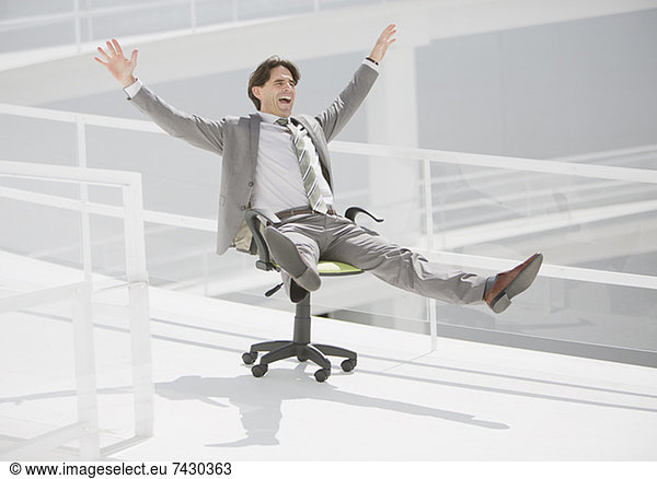 Carefree businessman sliding down walkway on office chair with wheels