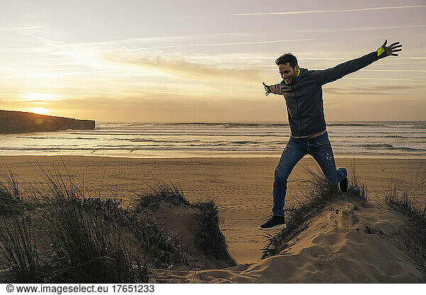 Carefree man jumping from sand dune with arms outstretched at beach