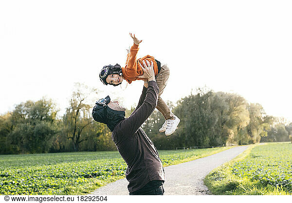 Carefree man lifting son wearing flying goggles in nature
