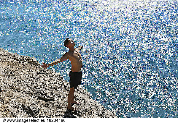 Carefree man with arms outstretched standing on rock by sea