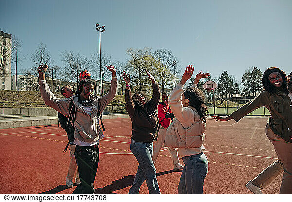 Carefree multiracial friends with arms raised dancing together at sports court on sunny day