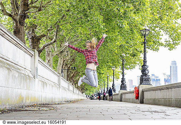 Carefree woman wearing checked shirt with arms raised jumping on footpath against trees