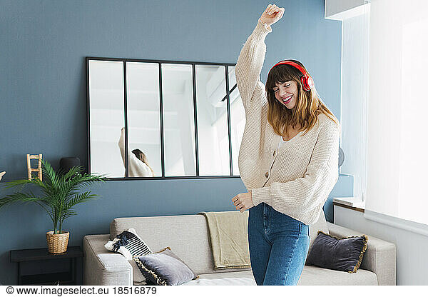 Carefree woman wearing headphones dancing with hand raised in living room at home