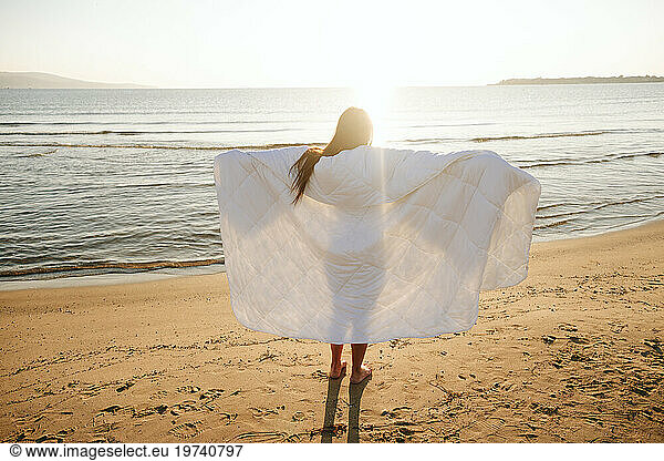 Carefree woman with arms outstretched wrapped in blanket at beach