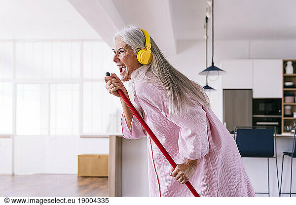 Carefree woman with yellow headphones listening to music holding broom at home