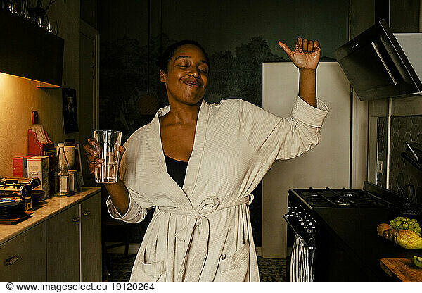 Carefree young woman holding drinking glass while dancing in kitchen at home
