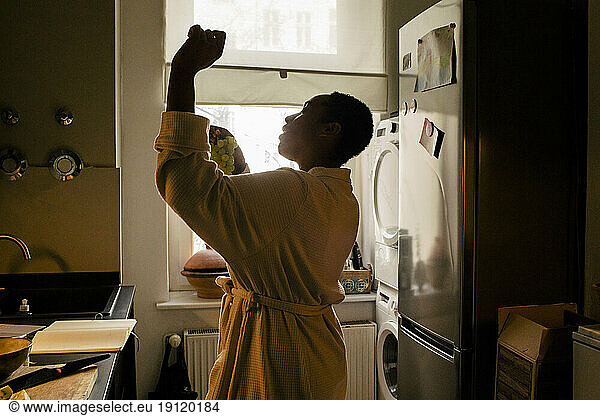 Carefree young woman holding grapes dancing in kitchen at home
