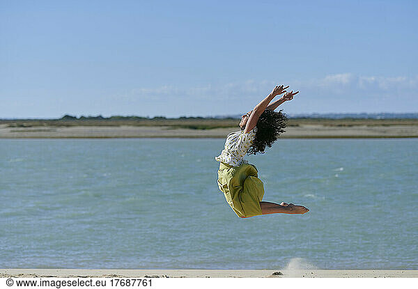 Carefree young woman jumping with arms raised at beach