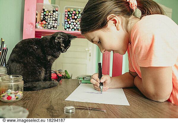 Cat watching girl drawing picture on paper at home