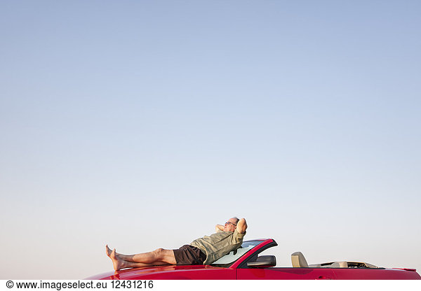 Caucasian male relaxing on the hood of his convertible sports car.
