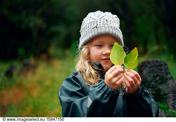 Charming girl in warm hat holding green leaves in forest