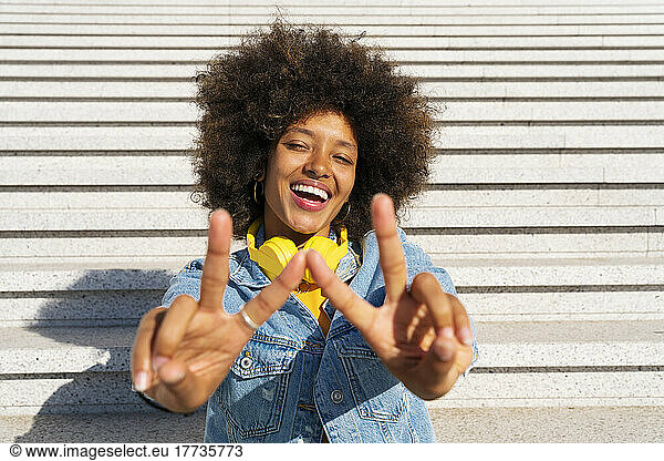 Cheerful Afro woman gesturing peace sign sitting on steps