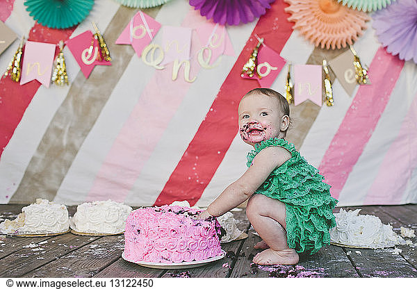 Cheerful baby girl looking away while eating birthday cake on floorboard during party