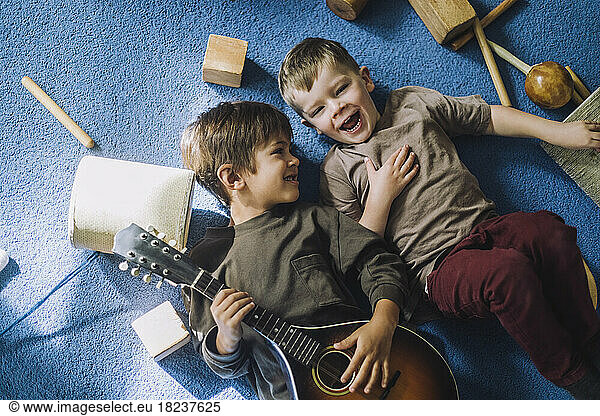 Cheerful boy lying down with male classmate holding guitar in kindergarten