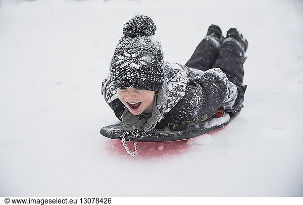 Cheerful boy tobogganing on snow during snowfall