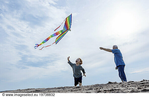 Cheerful boy with grandfather flying kite under sky at beach