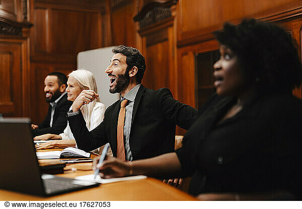 Cheerful businessman with mouth open sitting amidst colleagues in conference meeting at board room
