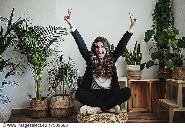 Cheerful businesswoman showing peace sign sitting cross-legged at office