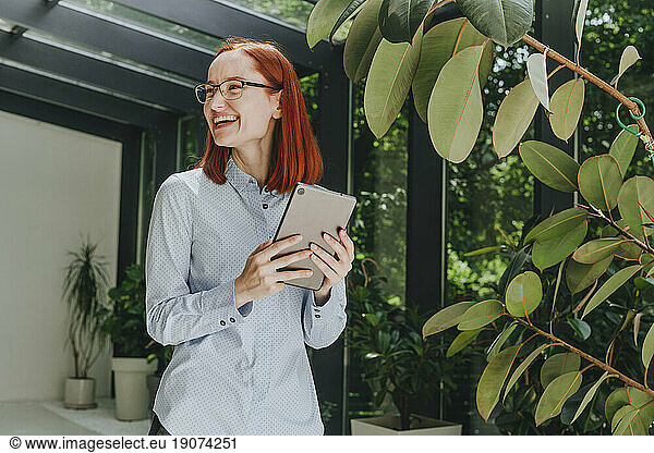 Cheerful businesswoman standing with tablet PC by plant in office