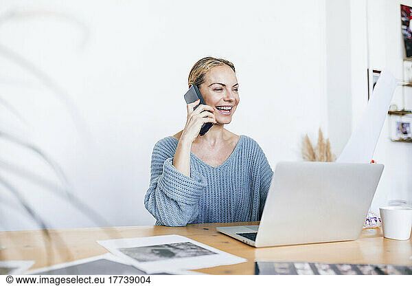 Cheerful businesswoman talking on smart phone sitting with laptop at home