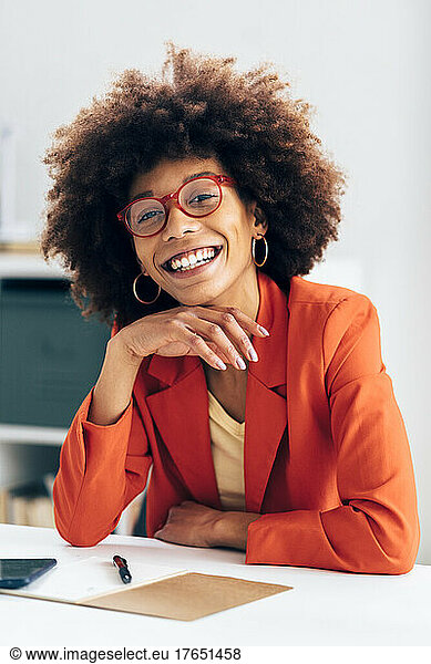 Cheerful businesswoman wearing eyeglasses sitting with hand on chin at desk