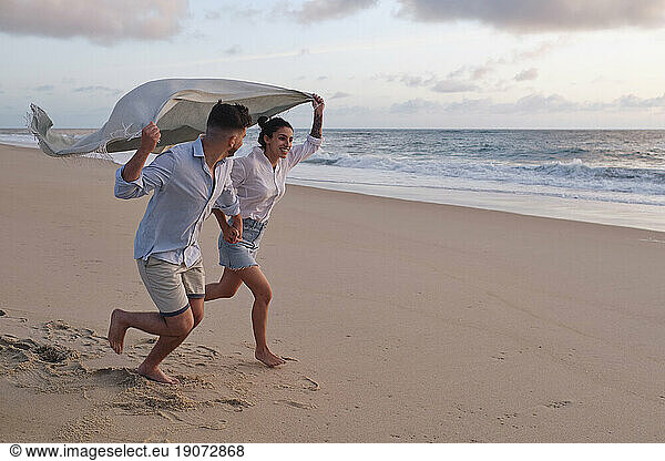 Cheerful couple running together holding blanket at beach