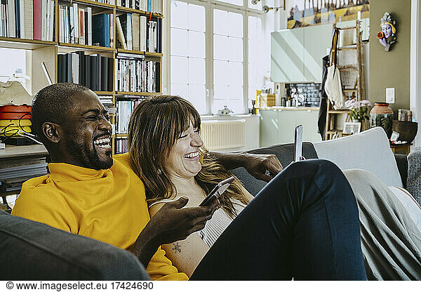 Cheerful couple sitting together with mobile phones on sofa in living room at home