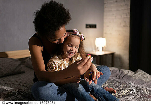 Cheerful ethnic mother and daughter lying in bed before sleep