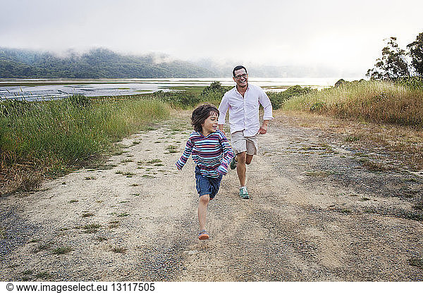 Cheerful father and son running on dirt road against cloudy sky
