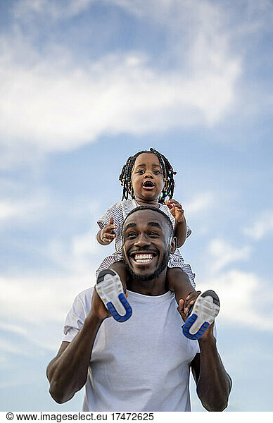 Cheerful father carrying daughter on shoulders under sky