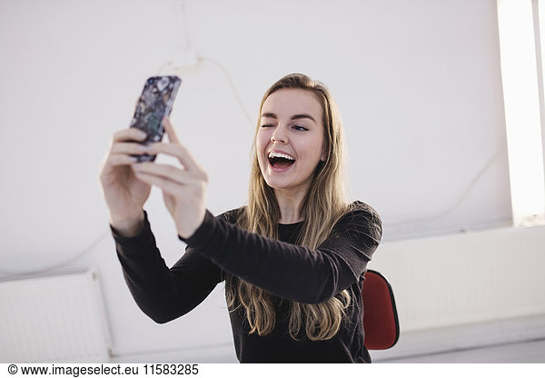 Cheerful female blogger winking while taking selfie through smart phone in office