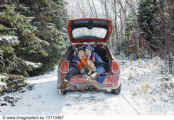 Cheerful female friends sitting in car trunk at Algonquin Provincial Park during winter