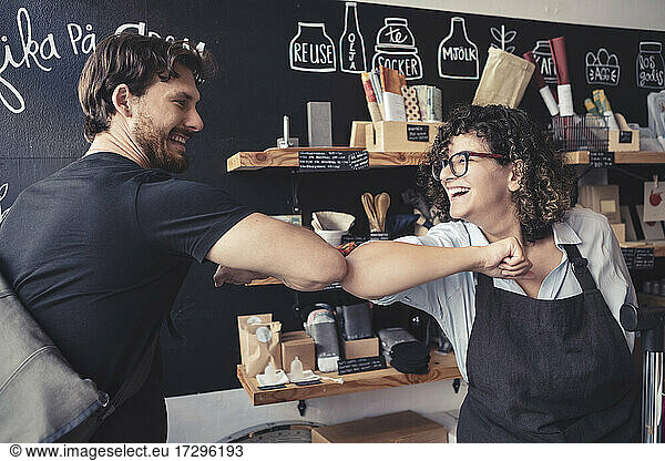 Cheerful female owner and male customer greeting with elbow bump at organic shop