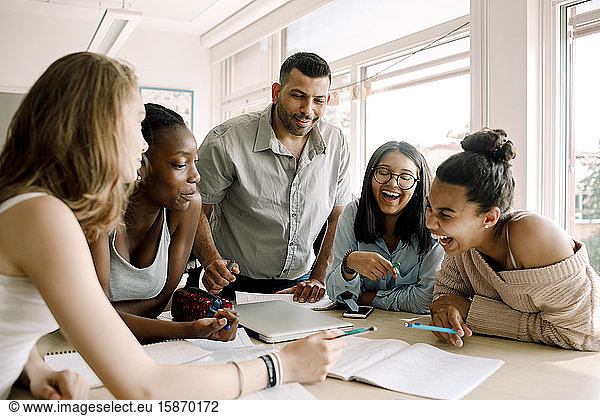 Cheerful female teenagers studying while professor standing by table in classroom