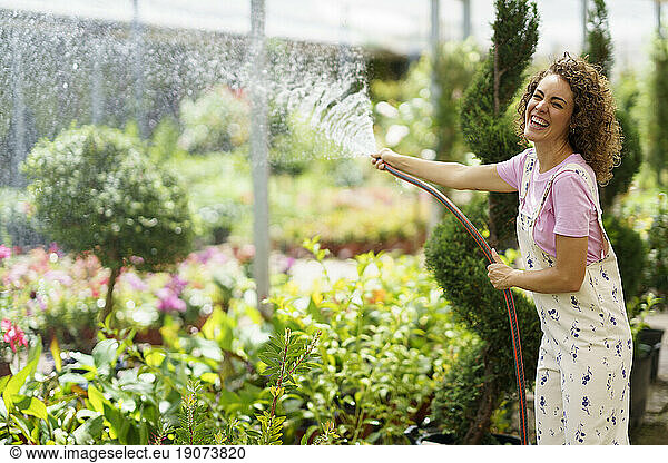 Cheerful florist watering plants with hose standing in nursery