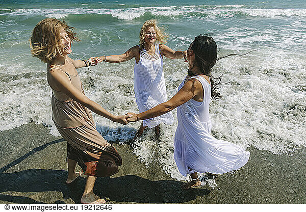 Cheerful friends holding hands and playing ring around the rosy near sea at beach