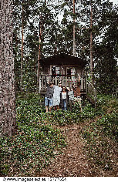 Cheerful friends with arms around standing against house in forest during vacation