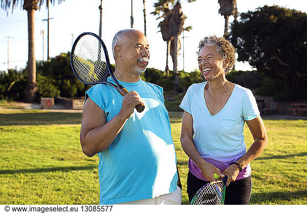 Cheerful friends with rackets talking while standing in park
