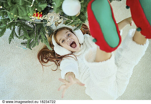 Cheerful girl lying on carpet by Christmas tree at home