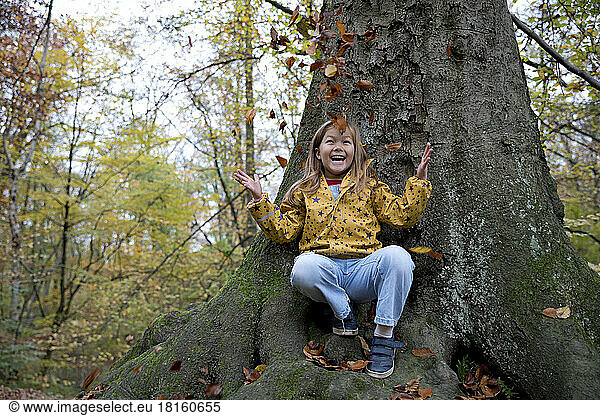 Cheerful girl playing with leaves sitting on tree trunk in forest