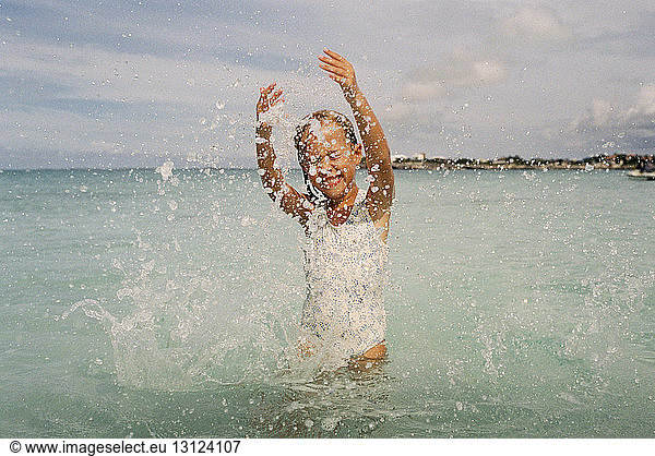 Cheerful girl splashing water in sea against cloudy sky