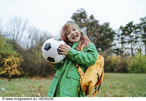 Cheerful girl standing with backpack and soccer ball at park
