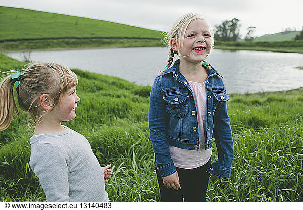 Cheerful girl standing with sister on grassy field by lake