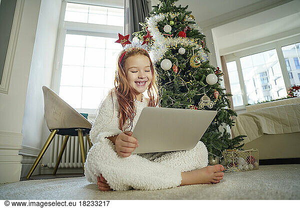 Cheerful girl using laptop sitting in front of Christmas tree at home