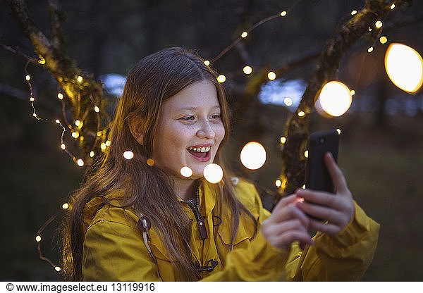 Cheerful girl using smart phone amidst illuminated string lights decoration at night