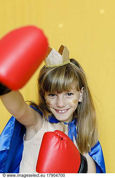 Cheerful girl wearing boxing gloves and crown against yellow background
