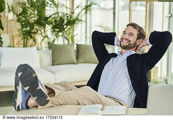 Cheerful male freelancer looking away sitting with feet up on table in home office