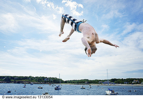 Cheerful man backflipping into sea against sky