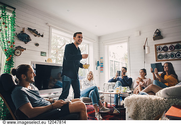 Cheerful man standing amidst young friends sitting in living room at home