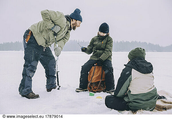 Cheerful man using ice auger while talking with backpack at frozen lake